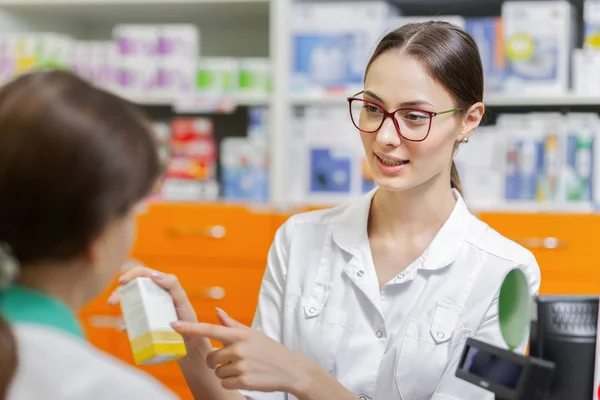 Una chica sonriente con cabello oscuro y gafas, con una bata de laboratorio, sugiere medicamentos a un visitante en la caja de una farmacia . —  Fotos de Stock