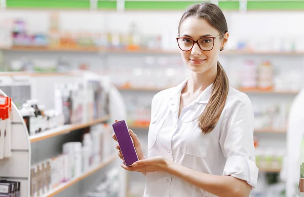 A smiling slim dark-haired lady with glasses, wearing a white coat, stands next to the shelf and shows some cosmetics in the modern pharmacy.