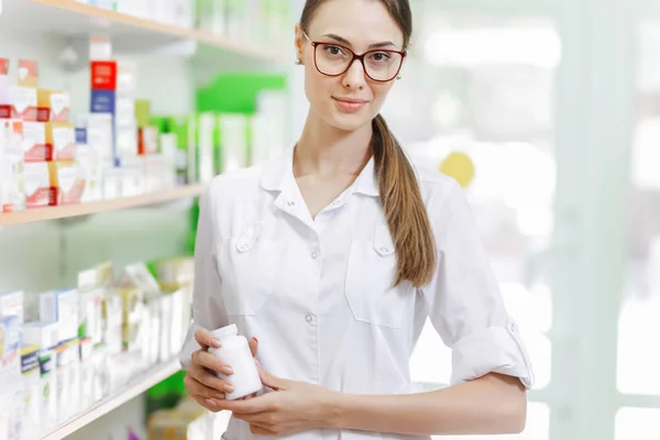 Una chica delgada y hermosa.Con una bata de laboratorio, muestra un frasco pequeño a la cámara en una farmacia moderna . — Foto de Stock