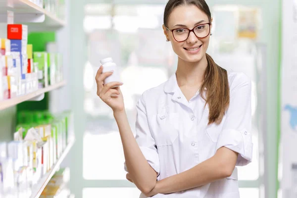 A youthful thin brown-haired lady with glasses,dressed in a lab coat,is holding a small jar in her right hand in a new pharmacy,