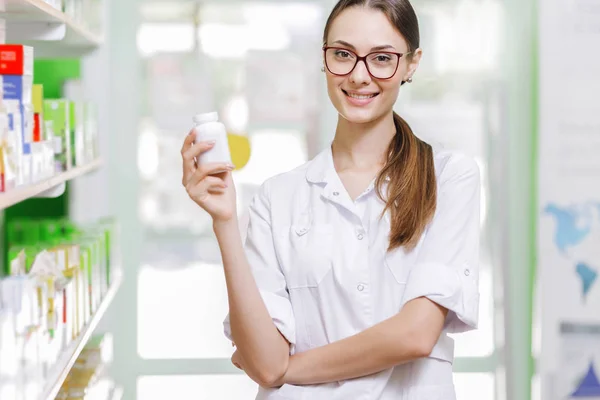 Una joven de pelo castaño delgado con gafas, vestida con una bata de laboratorio, sostiene un pequeño frasco en su mano derecha en una nueva farmacia , —  Fotos de Stock