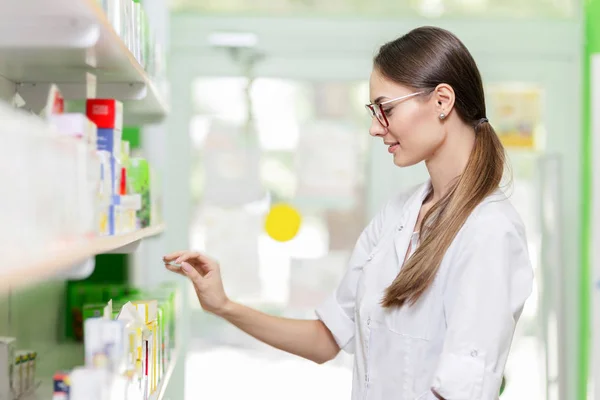A good looking slim girl with dark hair and glasses,wearing a lab coat,takes something from the shelf in a new pharmacy.