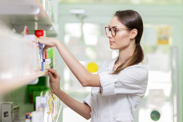 A cute thin lady with dark hair and glasses,wearing a lab coat,takes something from the shelf in an up-to-date  pharmacy.