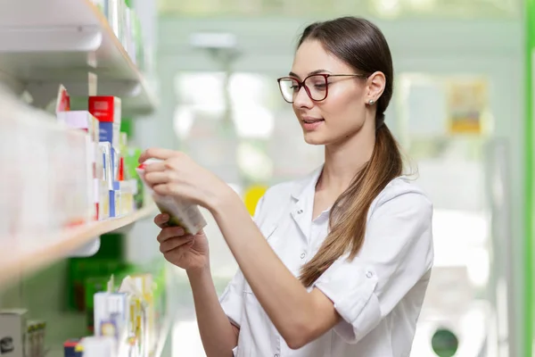 Una encantadora chica sonriente con cabello oscuro y gafas, con una bata de laboratorio, toma algo de la estantería en una farmacia moderna . —  Fotos de Stock