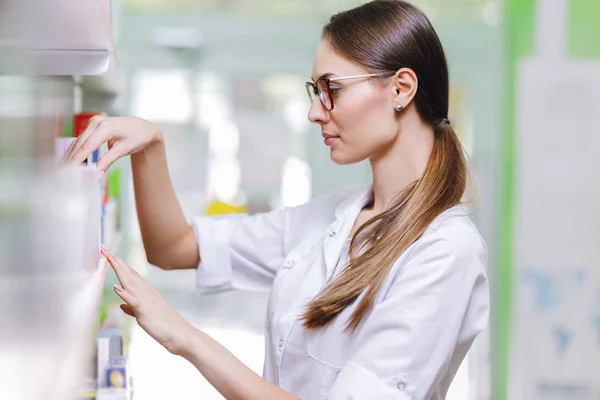 Una linda mujer delgada con cabello oscuro y gafas, con una bata de laboratorio, toma algo de la estantería en una farmacia actualizada . — Foto de Stock