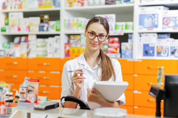 A beautiful thin dark-haired girl with glasses,dressed in a lab coat,writes notes in a notebook at the cash desk and looking at the camera in a modern pharmacy.