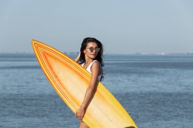 Güzel ince esmer kız güneş gözlüklü bir beyaz giyinmiş t-shirt kumlu plajda denize yakın yürüyüş ve sarı bir sörf tahtası onun elinde tutan