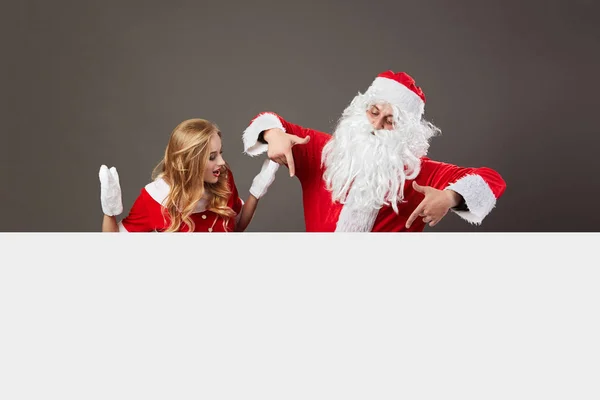 Санта Клаус і молодий красивою Місіс Клаус стояти за білому полотні і gesticulate на сірий фон. — стокове фото