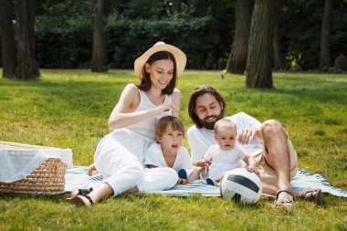 Yaz pikniği. İki çocuklu mutlu anne rahat kıyafetler yorgan yeşil çimenlerin üzerinde oturan beyaz giyinmiş.