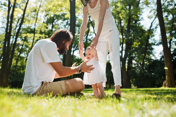 Urocze dziewczynki w białej sukni jej matka trzyma za rękę i stara się uczynić swoje pierwsze kroki w kierunku jej ojciec na zielony trawnik. — Zdjęcie stockowe