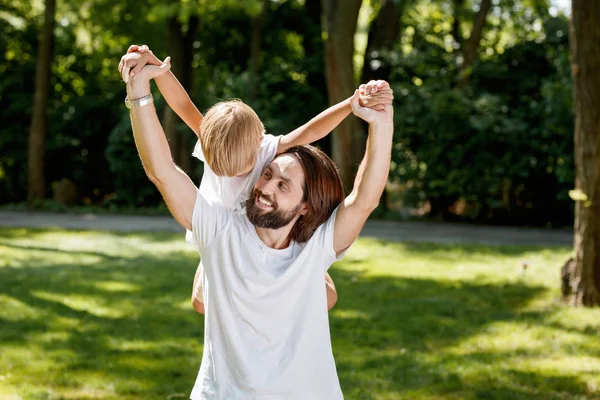 Homem bonito alegre dreesed na camisa branca do t está segurando seu filho pequeno em suas costas e rindo ao ar livre . — Fotografia de Stock
