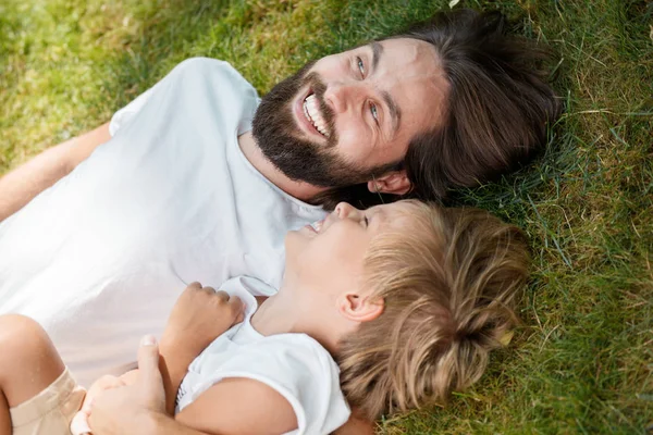 Gioioso padre e figlio sono sdraiati insieme sull'erba verde e ridono in una giornata di sole . — Foto Stock