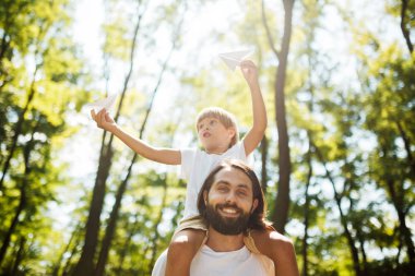 Yakışıklı baba ve küçük oğlu ormanda yürüyüş. Çocuk babasının omuzlarında oturan ve elinde bir kağıt uçak tutar.