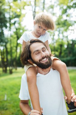 Baba ve oğlu ormanda yürüyüş. Çocuk babasının omuzlarında oturan beyaz tişört giymiş.