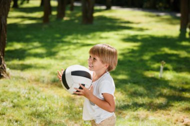 T gömlek ve bej şort giyen mutlu sarışın çocuk kollarını ve gülümseme futbol topu tutan çimenlerin üzerinde duruyor.