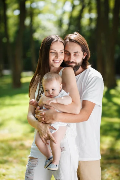 Junge hübsche brünette Mann umarmt sanft seine schöne Frau und kleine Tochter im Park an einem sonnigen Tag — Stockfoto