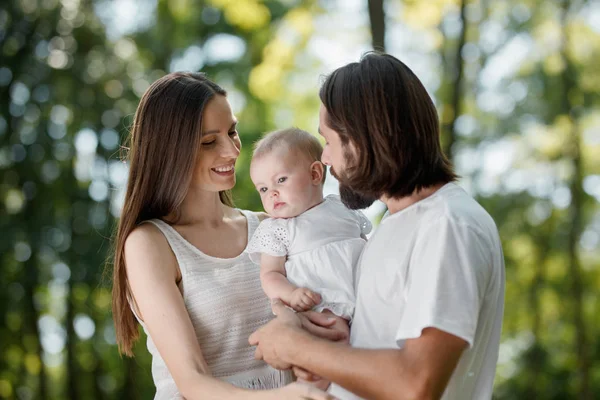 Nette junge Eltern in weißen Kleidern halten ihre charmante kleine Tochter in den Armen und lächeln zusammen. — Stockfoto