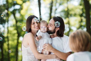 Gülümseyen aile beyaz giysiler giymiş küçük kızı kollarında tutarak ve Sabun kabarcık arayanlar.