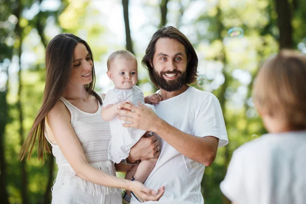 Faszinierende junge Eltern in weißen Kleidern halten ihre Tochter in den Armen, schauen den Sohn an und lächeln. — Stockfoto