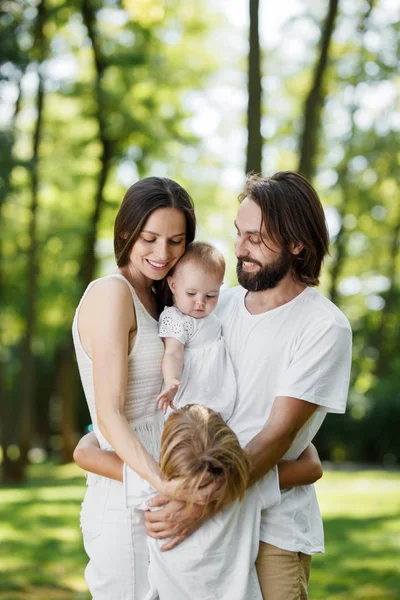 Jolie et amicale famille se repose dans le parc. Papa et maman tiennent leur fille dans les bras et embrassent leur fils . — Photo