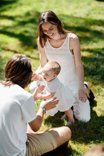 Uśmiechający się rodzice ubrani w białe szaty uczy ich córeczka jak zrobić swoje pierwsze kroki na trawniku. — Zdjęcie stockowe