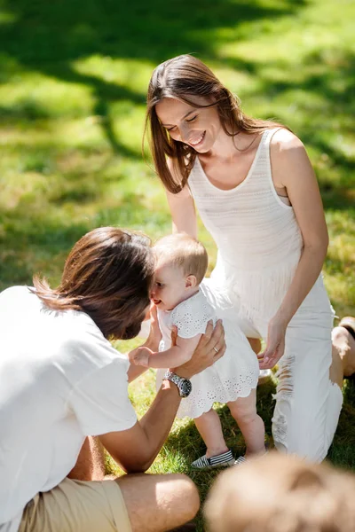 Dziewczynka w białej sukni starać się jej pierwsze kroki na trawniku, podczas gdy jej rodzice uśmiech i pomóc jej. — Zdjęcie stockowe