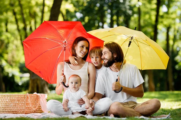 Se cachant du soleil sous de grands parapluies rouges et jaunes mère, père et leurs enfants sont assis sur la couverture et parlent . — Photo