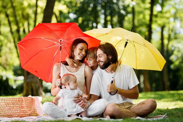 Sentado bajo paraguas de color rojo brillante y amarillo familia vestida con la ropa blanca tienen un descanso al aire libre —  Fotos de Stock