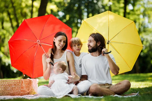 Enfants drôles avec maman et papa assis sur la couverture sous les grands parapluies rouges et jaunes les couvrant du soleil . — Photo