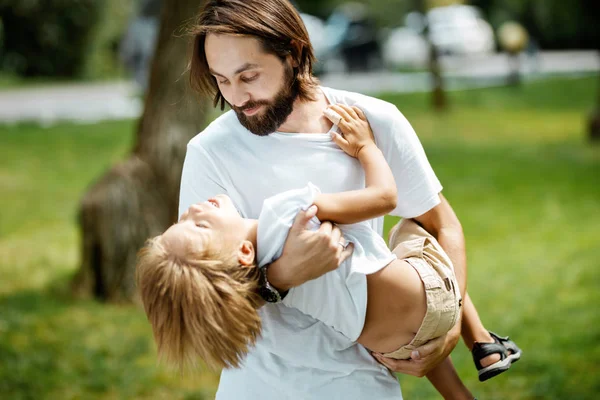 Handsome dark-haired father with beard dressed in the white t-shirt is holding in the arms his little son and watching at him on a summer day in the park.