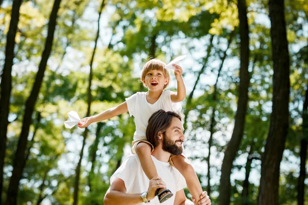 Ragazzo felice con suo padre vestito con le magliette bianche che passeggiano nel parco. Ragazzo è seduto sulle spalle dei padri e tiene aerei di carta nelle sue mani . — Foto Stock