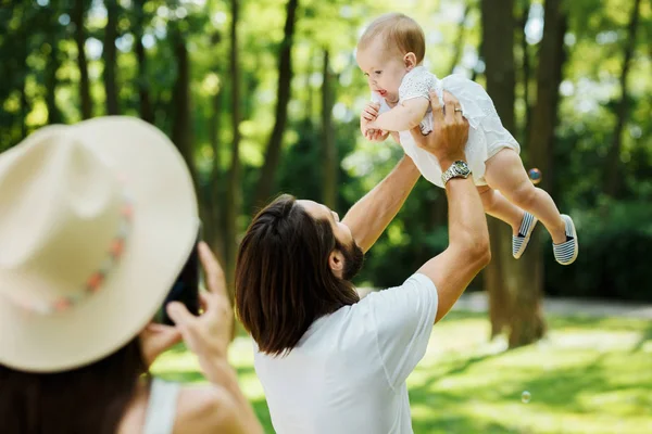 Junge Frau mit Hut macht ein Foto von ihrem Mann, der ihre kleine charmante Tochter an einem Sommertag im Park in den Armen hält. — Stockfoto