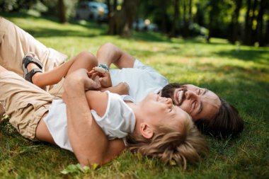 Bir sakallı koyu saçlı adam yatıyor taze yeşil bir çimenlikte küçük çocuk ile hava ve gülüyor.