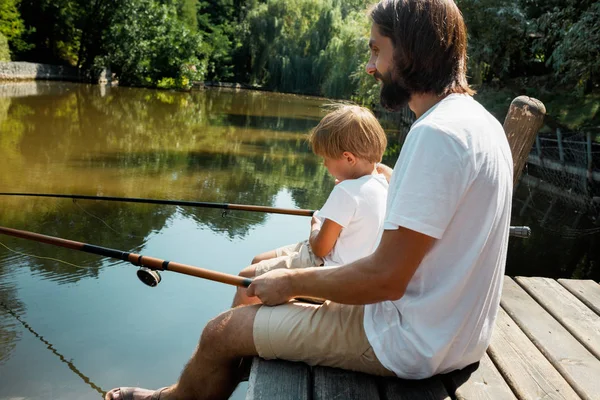 Der junge dunkelhaarige Vater sitzt mit seinem kleinen Sohn in weißen T-Shirts mit Angelruten auf dem Holzsteg und angelt an einem warmen Tag.. — Stockfoto