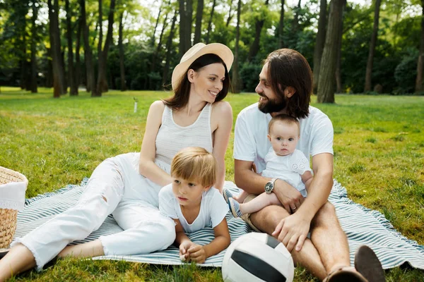 Belle jeune famille vêtue de vêtements blancs est assise sur une couverture sur la pelouse par une chaude journée ensoleillée dans le parc . — Photo