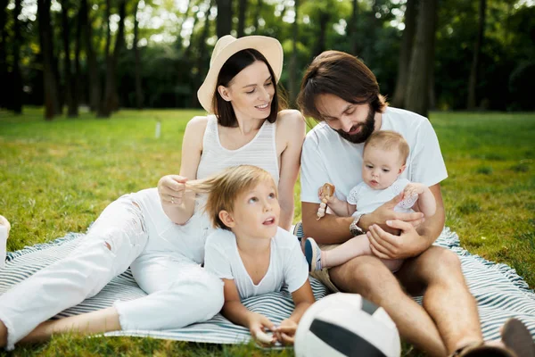 Cheerful parents with two kids dressed in white clothes is sitting on a striped blanket on the lawn on a warm sunny day in the park. . .