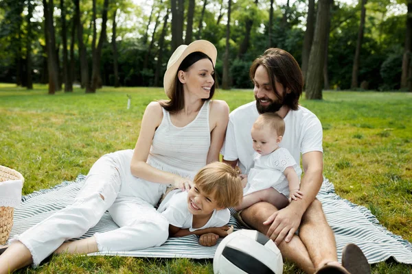 Des parents joyeux avec deux enfants vêtus de vêtements blancs sont assis sur une couverture rayée sur la pelouse par une chaude journée ensoleillée dans le parc . — Photo