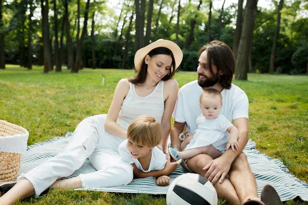 Des parents joyeux avec deux enfants vêtus de vêtements blancs sont assis sur une couverture rayée sur la pelouse par une chaude journée ensoleillée dans le parc . — Photo