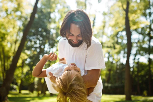 Przystojny szatyn ojca z brodą, ubrany w biały t-shirt jest trzymając w ramionach swojego małego synka i oglądając go w letni dzień w parku. — Zdjęcie stockowe