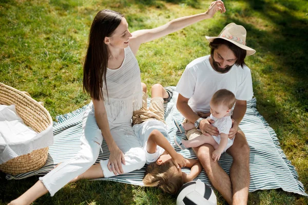 Journée chaude et ensoleillée dans le parc. Élégante jeune famille vêtue de vêtements décontractés blancs est assis sur une couverture sur l'herbe verte . — Photo