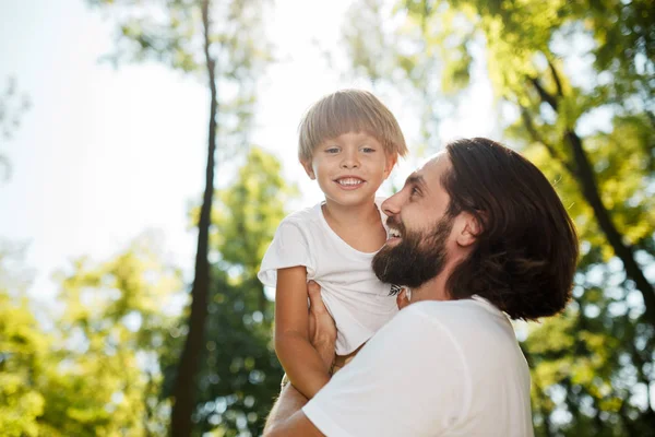 Przystojny szatyn ojca z brodą, ubrany w biały t-shirt trzyma w ramionach jego synek, oglądając go i uśmiechając się w parku. — Zdjęcie stockowe