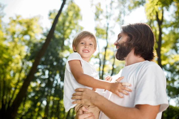 Handsome dark-haired father with beard dressed in the white t-shirt is holding in the arms his little son and watching at him on a summer day in the park.