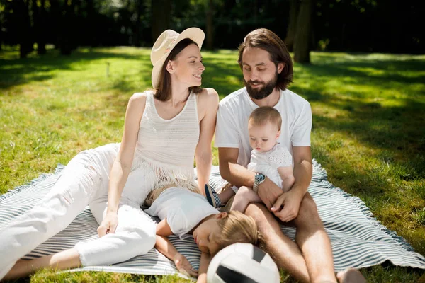 Journée chaude et ensoleillée dans le parc. Élégante jeune famille vêtue de vêtements décontractés blancs est assis sur une couverture sur l'herbe verte . — Photo
