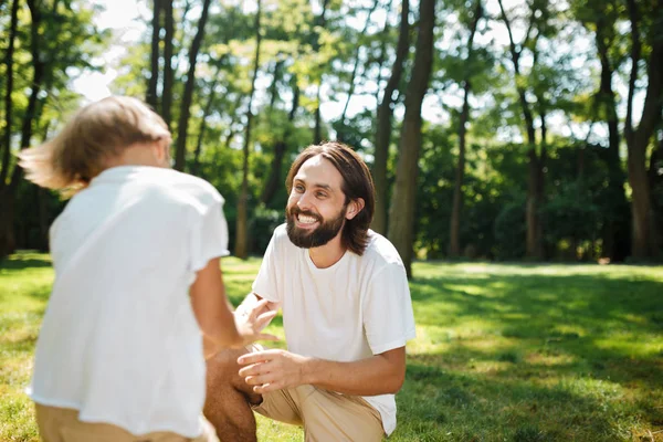 Papà dai capelli scuri e suo figlio vestito con magliette bianche si divertono sul prato verde del parco in una giornata di sole — Foto Stock