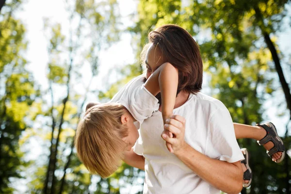 Dark-haired father with beard dressed in the white t-shirt is holding in the arms his blond son and smiling on a summer day in the park.