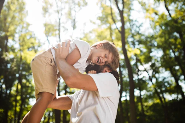 Padre dai capelli scuri con la barba vestita con la t-shirt bianca tiene tra le braccia suo figlio biondo e sorride in una giornata estiva nel parco . — Foto Stock