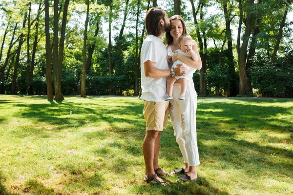 Pai de cabelos escuros beija sua bela esposa segurando uma pequena filha em um vestido branco em um parque em um dia ensolarado . — Fotografia de Stock