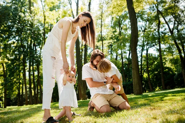 Feliz jovem mãe de cabelos escuros e pai vestido com roupas brancas estão passando o dia de verão com as crianças em um gramado no parque . — Fotografia de Stock