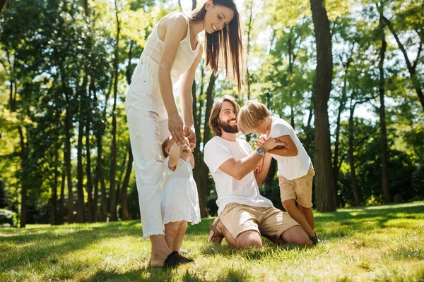 Szczęśliwa młoda Ciemnowłosa matka i ojciec, ubranych w białe szaty spędzają letni dzień z dziećmi na trawnik w parku. — Zdjęcie stockowe