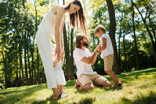 Felice giovane madre dai capelli scuri e padre vestito con abiti bianchi stanno trascorrendo la giornata estiva con i bambini su un prato nel parco . — Foto Stock